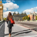 A woman with a red bag walks toward the Palace of Westminster on a bright, clear day.