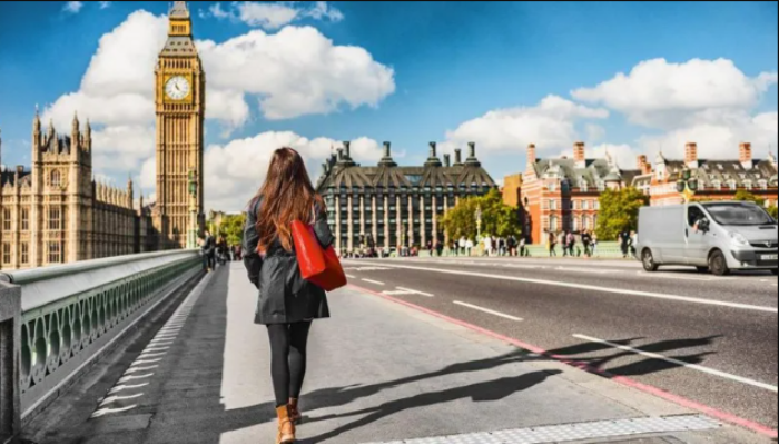 A woman with a red bag walks toward the Palace of Westminster on a bright, clear day.